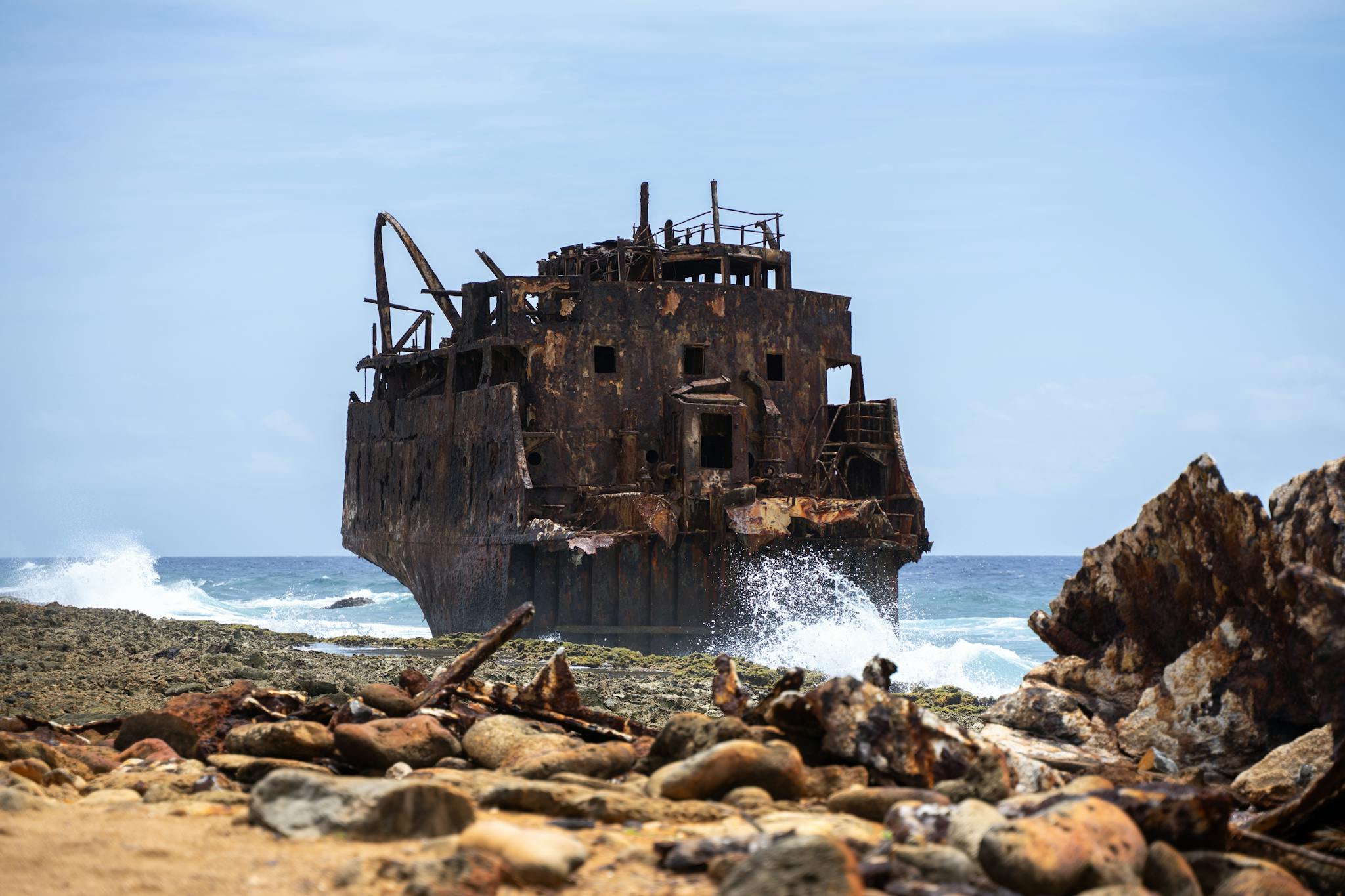 A rusting shipwreck on Curaçao's rocky coast with waves crashing, under a clear blue sky.